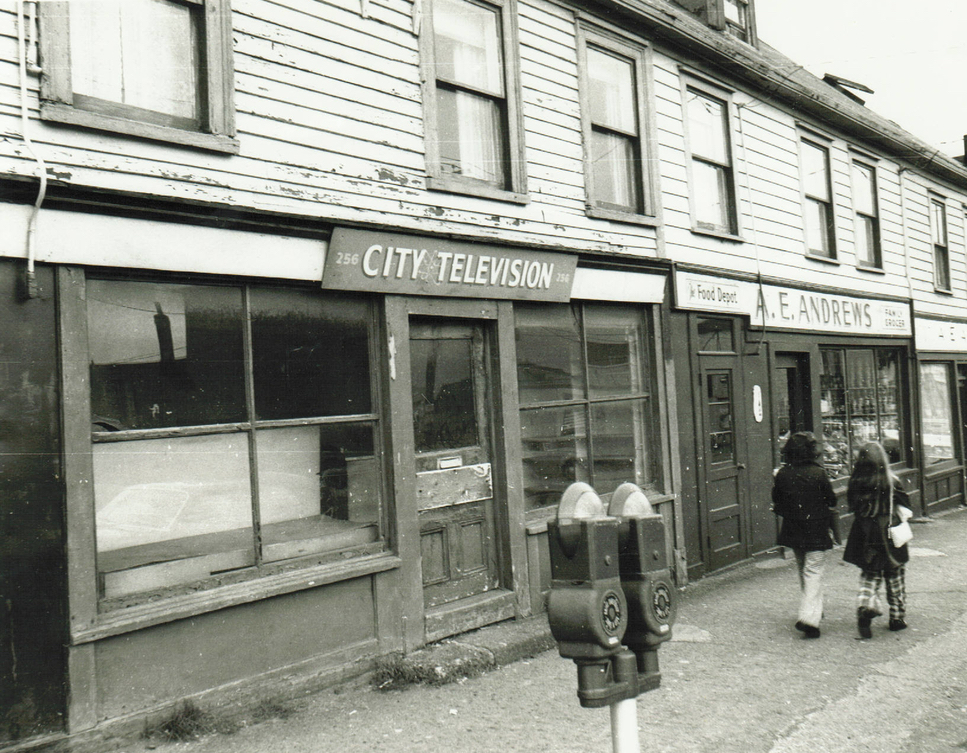 Paysage de rue en noir et blanc. Des maisons en rangée portent les enseignes City Television et A.E. Andrews. Deux femmes marchent sur le trottoir devant les bâtiments, vêtues de manteaux et de pantalons.