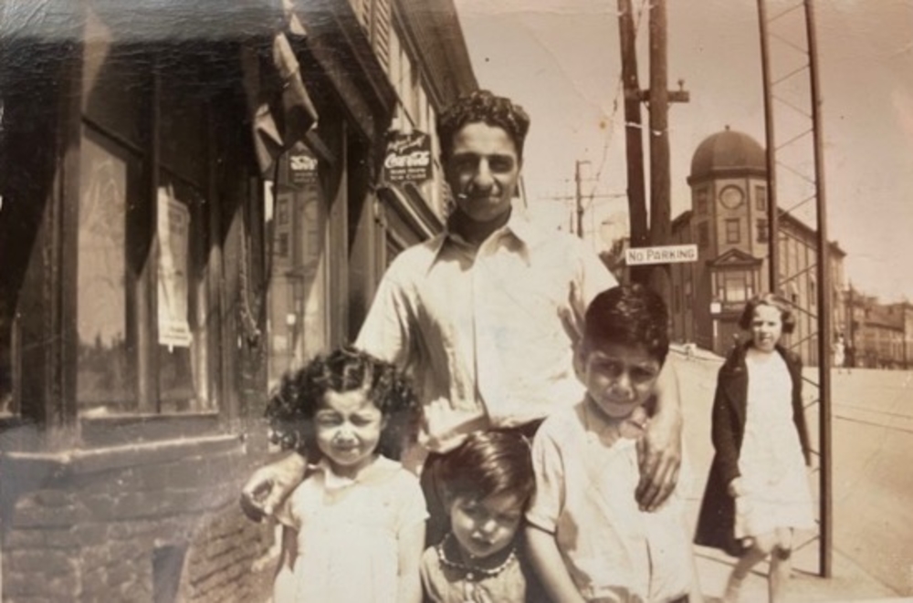 Photo d’extérieur en sépia prise sur le trottoir devant des magasins. Un homme se tient au centre avec une fille et deux garçons devant lui. Une autre jeune femme se tient à l’arrière-plan, à droite, vêtue d’une robe et d’un manteau.