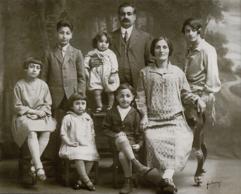 Portrait en noir et blanc d’une famille sur fond de forêt. De gauche à droite : une fille assise en robe et bas de couleur claire, un adolescent debout en costume, une fillette assise en robe et bas de couleur claire, un tout-petit debout en vêtements clairs, un homme en costume sombre avec sa main droite sur le tout-petit, un petit garçon assis en costume de marin sombre, une femme assise en robe claire, et une jeune femme en tenue claire. Le nom Holloway est signé dans le coin inférieur droit.