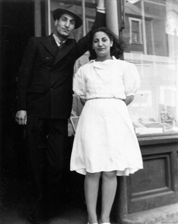 Portrait en noir et blanc. Un homme vêtu d’un costume sombre et d’un chapeau mou, portant une moustache sombre, se tient sur une marche avec une femme plus jeune aux cheveux foncés, vêtue d’une robe blanche et portant des chaussures blanches. L’homme est à gauche, la femme à droite. Les deux se tiennent sur le perron devant un magasin. La fenêtre derrière eux reflète les bâtiments situés de l’autre côté de la rue.