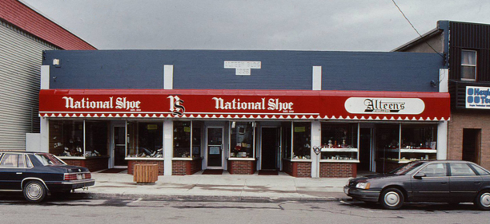 Photo extérieure en couleurs. Bâtiment en briques avec des enseignes rouges sur lesquelles on peut lire National Shoe; NS; National Shoe; Alteen’s. Au-dessus de l’enseigne, le bâtiment est peint en bleu. Au milieu, on peut lire Alteen Bldg 1953. Deux voitures sont stationnées à l’avant.