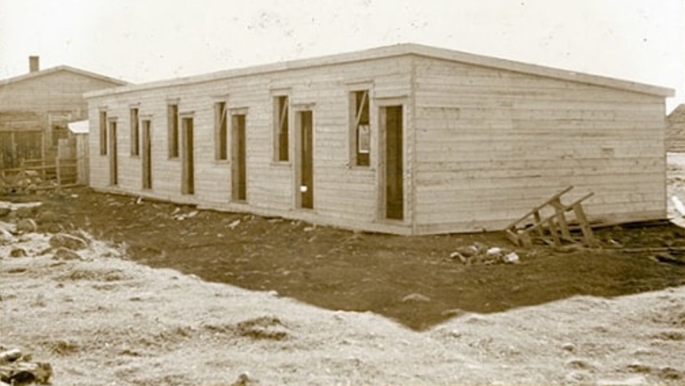 Photo d’extérieur en sépia. Bâtiment rectangulaire en construction. Six fenêtres et six portes sont visibles. Le bâtiment semble de couleur claire et projette une ombre sur le gravier. Un escabeau se trouve sur le côté droit du bâtiment. Un autre bâtiment se trouve à côté de celui-ci, sur le côté gauche.