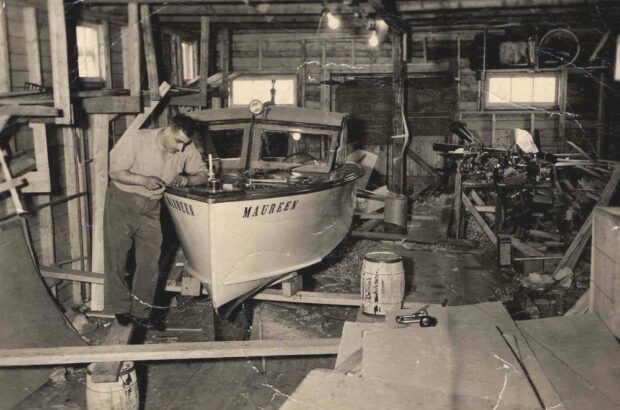 Photo d’intérieur en noir et blanc. Homme travaillant à un bateau dans un hangar ou une dépendance en bois avec cinq fenêtres. Le bateau se trouve sur le côté gauche de l’image et est posé sur des planches de bois et des barils. Le bateau comporte des couleurs claires et foncées et est muni d’un toit. Le mot Maureen est écrit de part et d’autre de la proue du bateau. Il y a des lumières fixées au centre du plafond. Du bois et des outils divers se trouvent sur le côté droit de l’image.