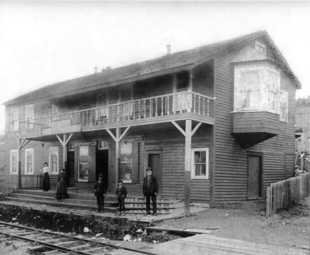 Photo d’extérieur en noir et blanc. Grand bâtiment de deux étages en arrière-plan avec un balcon au centre et une grande baie vitrée à droite. Deux femmes en jupe et en robe, deux jeunes garçons et un homme plus âgé en costume se tiennent sur les marches du bâtiment. Il y a une clôture autour du bâtiment, et devant les marches se trouvent des rails de chemin de fer.