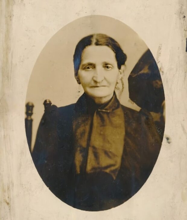 Portrait sépia d’une femme aux cheveux et aux yeux foncés portant des vêtements sombres. La femme a les cheveux relevés et est assise sur une chaise. Elle est au centre de la photo qui est de forme ovale, bien qu’il y ait quelqu’un à côté d’elle.