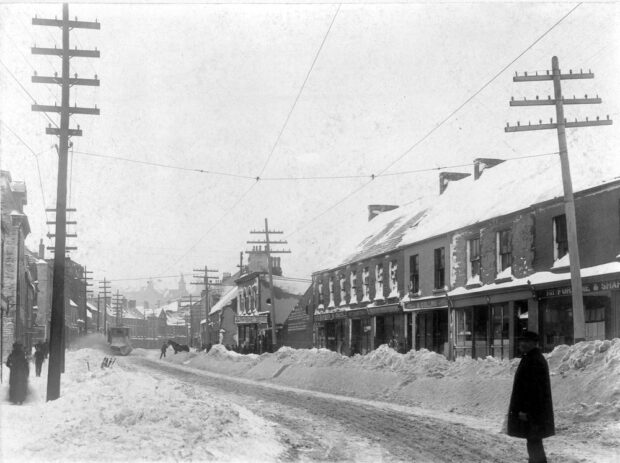 Paysage de rue en noir et blanc. Une rue et des trottoirs enneigés avec des maisons en rangée et des maisons attenantes en pierre et en briques ainsi que des poteaux électriques de chaque côté de la rue. Une douzaine de personnes se trouvent sur le trottoir. Il y a un homme dans la rue sur le côté droit à l’avant- plan de la photo. Un autre homme se trouve sur le côté droit de la rue, menant un cheval. Un chasse-neige déblaie le côté gauche de la rue.