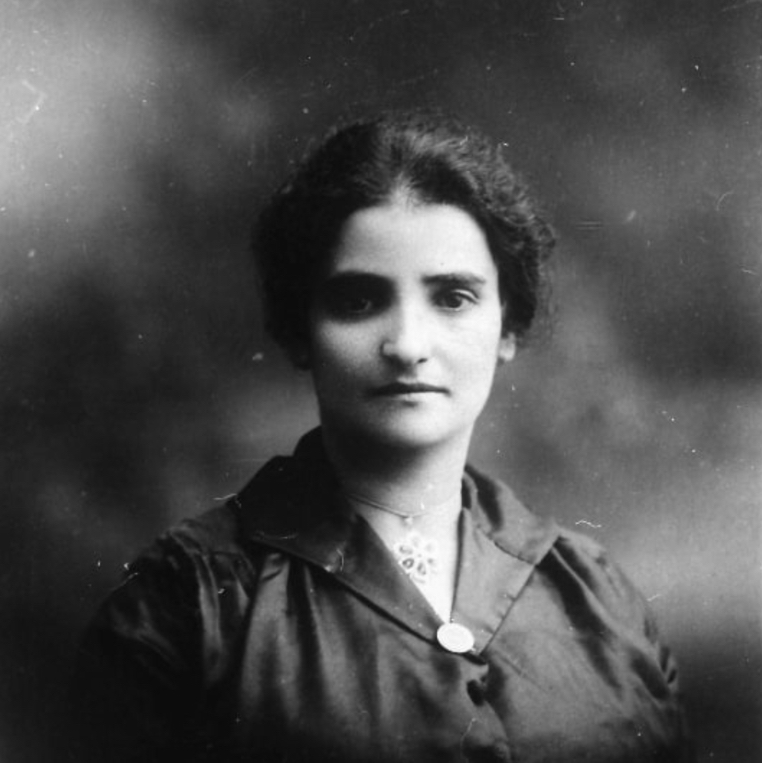 Portrait en noir et blanc d’une femme aux cheveux et aux yeux foncés, portant des vêtements sombres et un collier avec pendentif en forme de fleur et ayant les cheveux relevés.