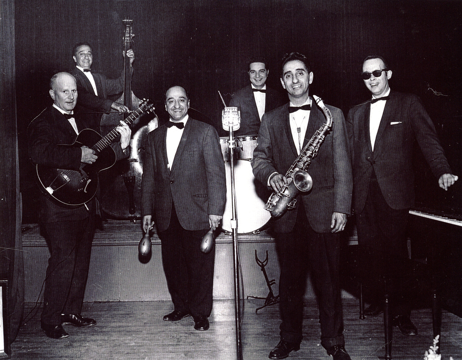 Portrait en noir et blanc. Groupe composé de six hommes habillés en costume, chacun tenant un instrument différent (de gauche à droite : guitare, violoncelle, maracas, batterie, saxophone et piano). Un microphone se trouve au centre de la photo. Le violoncelliste et le batteur sont respectivement debout et assis sur la scène, un peu plus haut que les quatre autres hommes qui se tiennent devant.