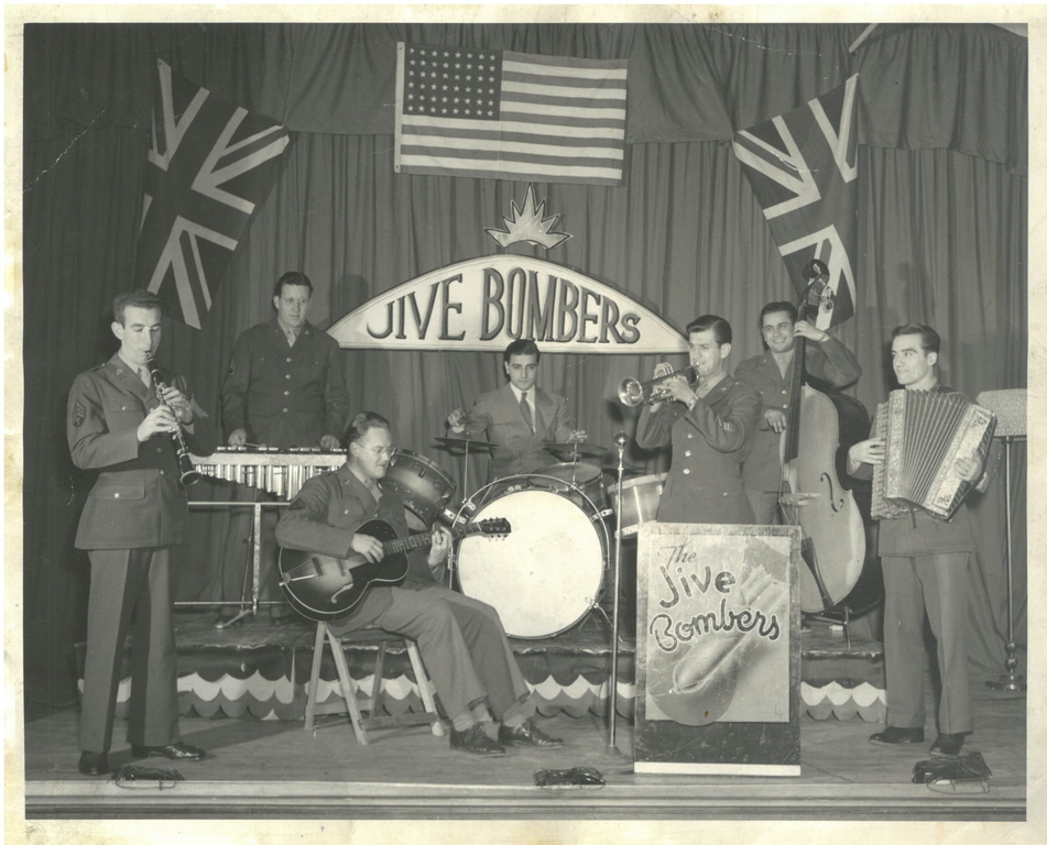 Photo d’intérieur en noir et blanc. Un groupe appelé The Jive Bombers compte trois hommes sur une scène à l’arrière et quatre hommes à l’avant. Les sept hommes portent des costumes ou des uniformes militaires et jouent d’un instrument. De gauche à droite : clarinette, vibraphone, guitare, batterie, trompette, violoncelle et accordéon. Trois drapeaux sont accrochés aux rideaux derrière et au-dessus des hommes : deux Union Jacks et un drapeau américain au centre. Sous le drapeau américain se trouve un panneau annonçant les Jive Bombers.