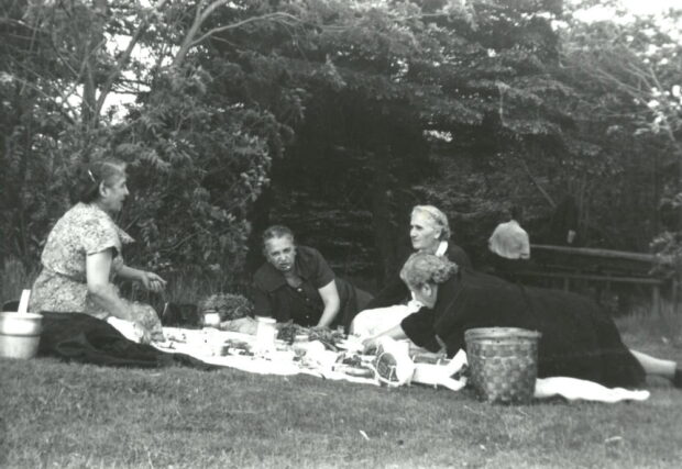 Photo d’extérieur en noir et blanc. Quatre femmes âgées pique-niquent dans un parc. Les femmes portent toutes des robes, et deux d’entre elles sont assises tandis que les deux autres sont allongées sur la couverture de pique-nique. Il y a divers aliments et deux paniers posés sur une couverture de couleur claire. Il y a des arbres à l’arrière-plan ainsi qu’une clôture en lisses, et un homme à l’arrière-plan à droite.