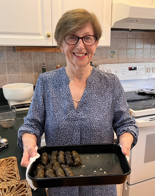 Une femme aux cheveux courts, portant des lunettes et un chemisier bleu et blanc, tient une casserole contenant neuf feuilles de vigne. Elle est debout dans une cuisine.