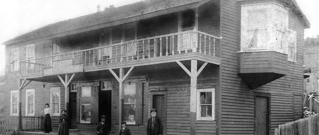 Photo d’extérieur en noir et blanc. Grand bâtiment de deux étages en arrière-plan avec un balcon au centre et une grande baie vitrée à droite. Deux femmes en jupe et en robe, deux jeunes garçons et un homme plus âgé en costume se tiennent sur les marches du bâtiment. Il y a une clôture autour du bâtiment, et devant les marches se trouvent des rails de chemin de fer.