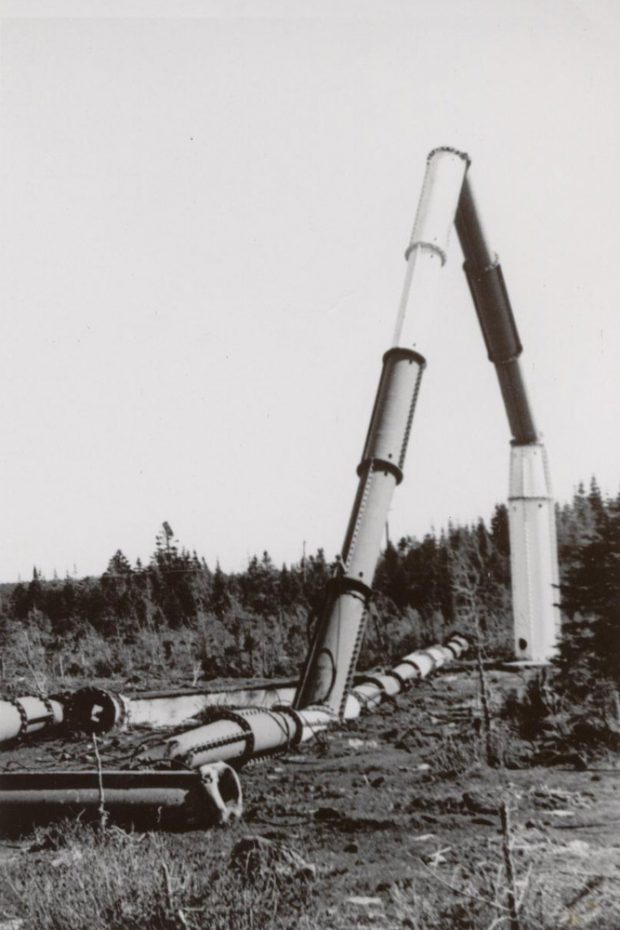 Sections de tubes métalliques ronds, certains debout reliés à une base, d'autres posés au sol, avec des sapins en arrière-plan.