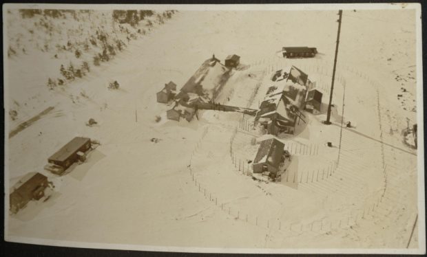 Vue aérienne de trois bâtiments et d'une antenne, entourés d'une clôture, et d'autres dépendances se trouvant à proximité. La neige recouvre le sol.