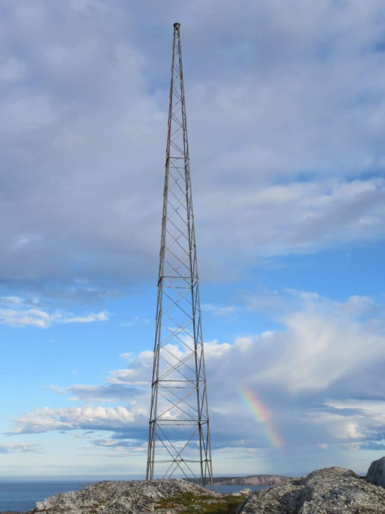 Grand pylône d'antenne s’élevant sur des rochers plats du littoral, un arc-en-ciel apparaissant derrière.