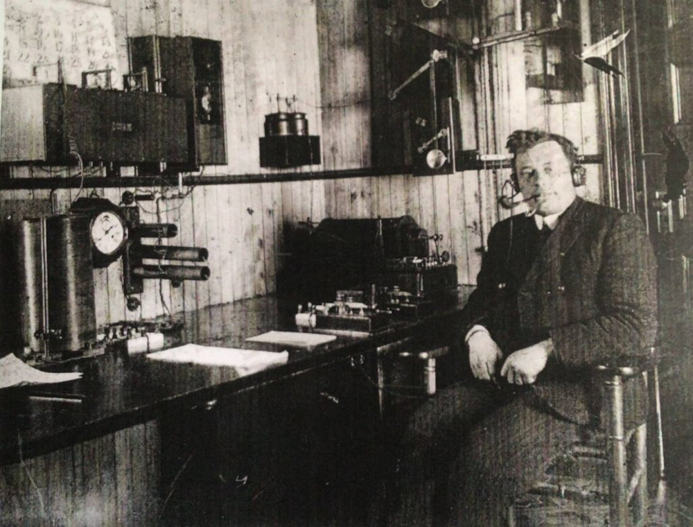 Un homme en complet est assis face à l’appareil-photo, à côté d'un bureau sur lequel sont posés du matériel de télégraphie sans fil et des piles de papier soigneusement ordonnées. Il porte un casque d'écoute et fume la pipe.