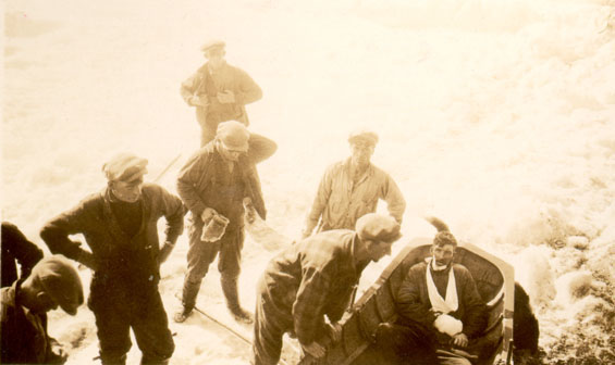 Sept hommes, vêtus de casquettes, de bottes et de manteaux, se tiennent sur une banquise. L'un d'eux se penche vers un petit bateau dans lequel se trouve un homme blessé. La main droite et la mâchoire du blessé ont des bandages.