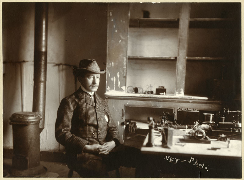 Un homme vêtu d'un veston à carreaux et d'un chapeau est assis à une table sur laquelle est posé du matériel de communication sans fil. Derrière lui se trouvent une armoire vide sans portes, ainsi qu’un poêle et un tuyau de poêle en métal.