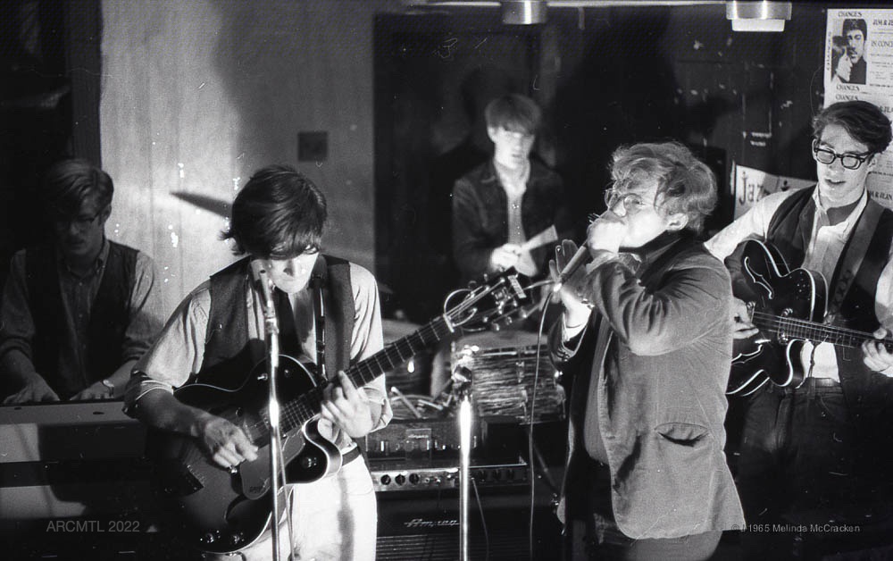 Photographie en noir et blanc de cinq musiciens se produisant dans un coin d'une pièce lambrissée. Deux guitaristes et un harmoniciste sont visibles au premier plan, un batteur et un joueur d'orgue sont visibles à l'arrière-plan.