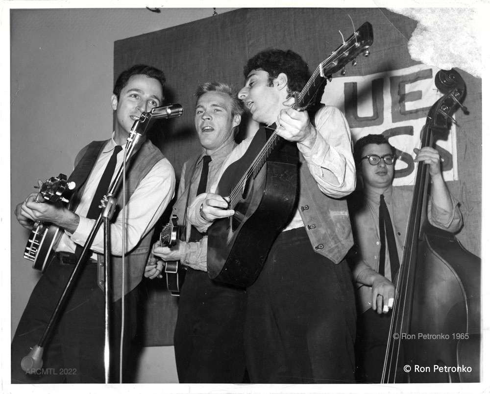 Photographie en noir et blanc de quatre hommes portant des gilets et des cravates, chantant et jouant de la guitare, de la mandoline et de la basse.