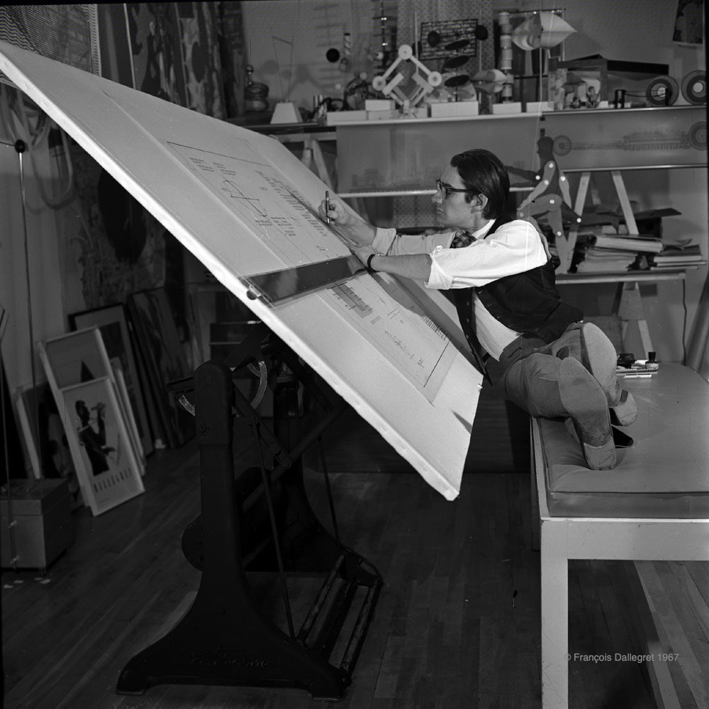 Photo en noir et blanc datant de 1966 de l'artiste François Dallegret assis sur un banc, les jambes relevées de côté, à sa grande table à dessin, en train de travailler sur des projets.