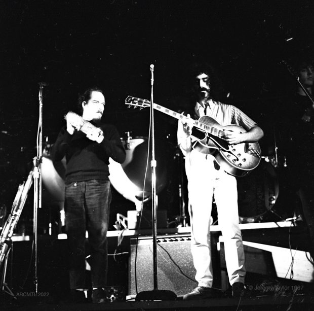 Photographie en noir et blanc de deux membres du groupe Mothers of Invention en concert au New Penelope, l'un joue du tambourin, l'autre est Frank Zappa avec une moustache et un bouc et de longs cheveux noirs, jouant de la guitare électrique.