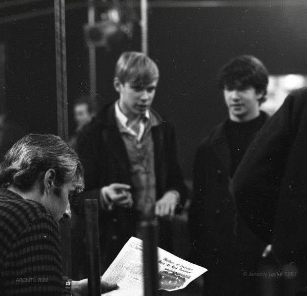 Photographie en noir et blanc de trois jeunes hommes, deux debout et un assis, dans le New Penelope. Au premier plan, l'un d'entre eux est assis sur un banc et lit un journal, à l'arrière-plan, les deux autres sont debout et le regardent. Le titre de l'article lu est Mothers Of Invention Now at New Penelope (Les mères de l'invention maintenant au New Penelope).