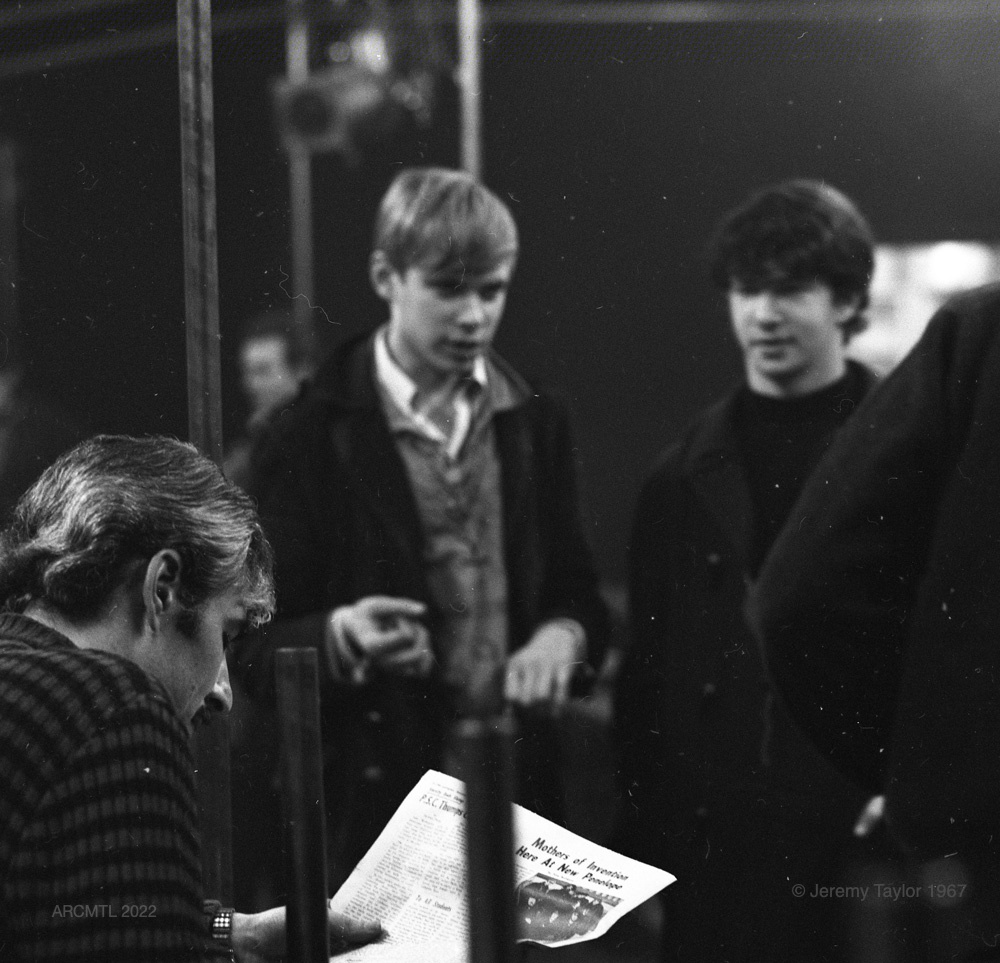 Photographie en noir et blanc de trois jeunes hommes, deux debout et un assis, dans le New Penelope. Au premier plan, l'un d'entre eux est assis sur un banc et lit un journal, à l'arrière-plan, les deux autres sont debout et le regardent. Le titre de l'article lu est "Mothers Of Invention Now at New Penelope" (Les mères de l'invention maintenant au New Penelope).