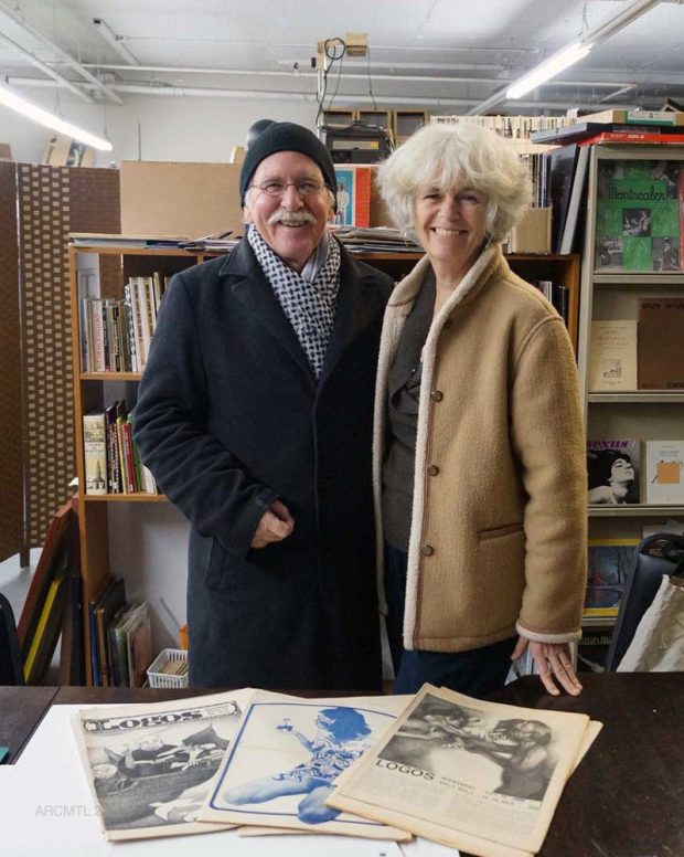 Photo d'un couple âgé portant des manteaux d'hiver et regardant l'appareil photo, debout devant les étagères d'une bibliothèque, avec trois exemplaires du journal Logos posés sur une table devant eux.