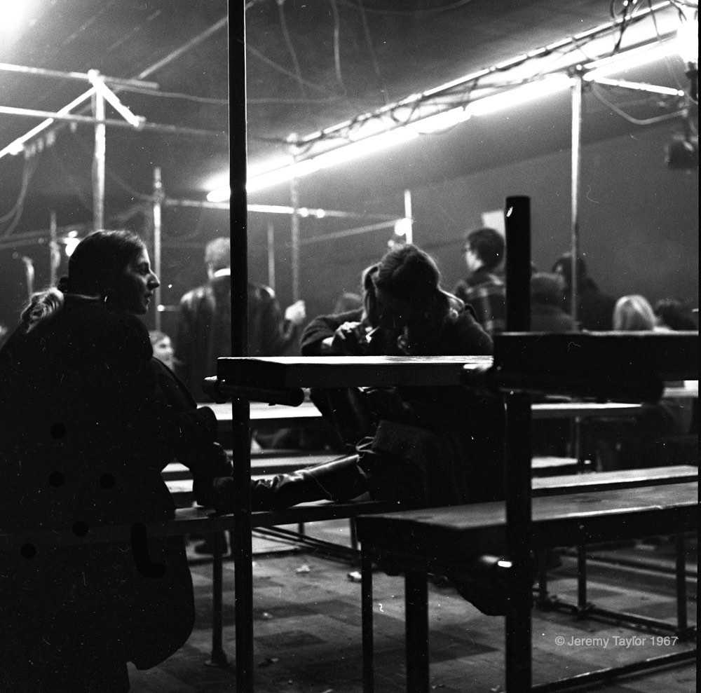 Photographie en noir et blanc de jeunes gens assis sur des bancs au New Penelope avec deux femmes en silhouette assises au premier plan, les pieds sur un banc. Des lumières vives sont suspendues à un échafaudage en haut de l'image.