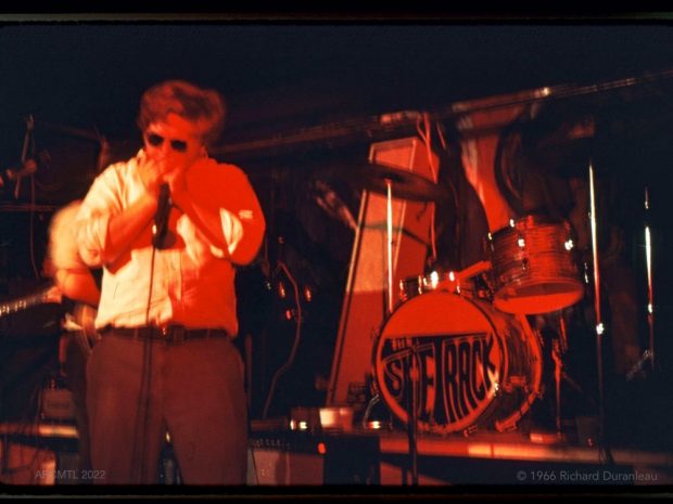 Sur une scène aux silhouettes rouges, un homme en chemise blanche, pantalon sombre et lunettes de soleil joue de l'harmonica dans un micro. À côté de lui, en arrière-plan, une batterie sur laquelle est inscrit le nom du groupe, The Sidetrack.