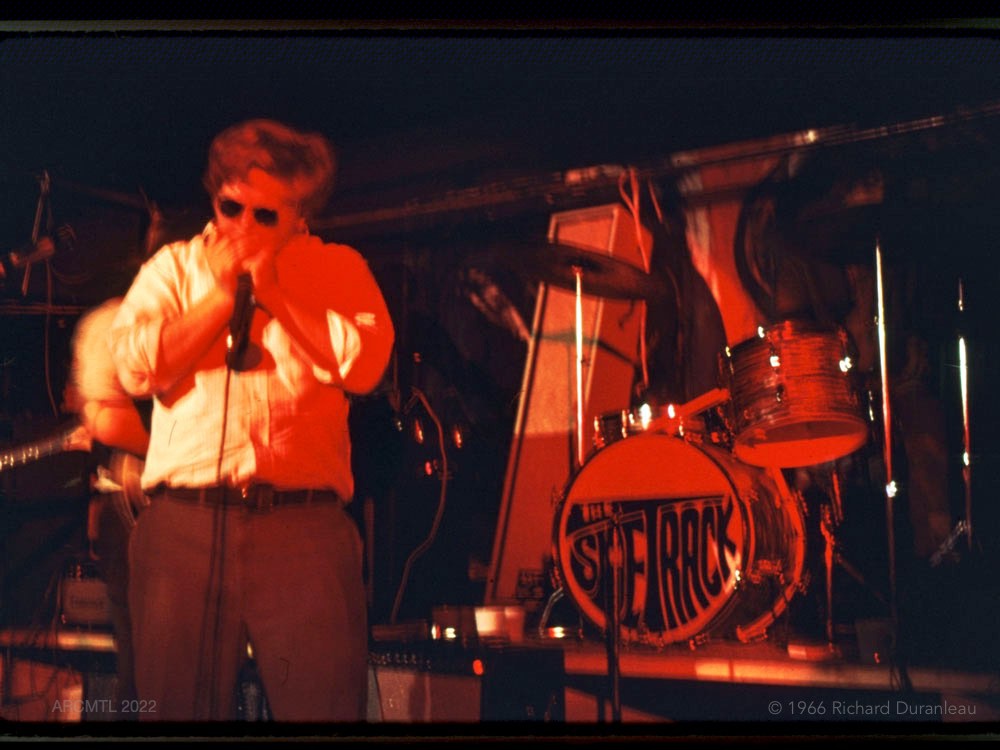 Sur une scène aux silhouettes rouges, un homme en chemise blanche, pantalon sombre et lunettes de soleil joue de l'harmonica dans un micro. À côté de lui, en arrière-plan, une batterie sur laquelle est inscrit le nom du groupe, "The Sidetrack".