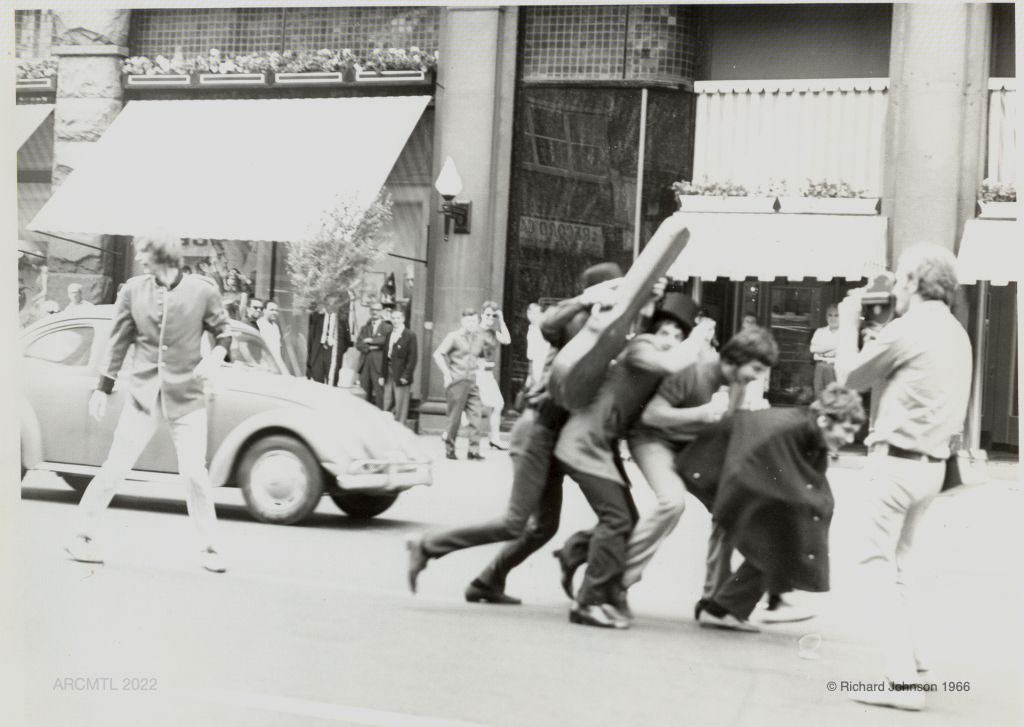 Photographie en noir et blanc d'un groupe de jeunes hommes souriants et pressés au milieu d'une rue commerçante, portant des chapeaux haut de forme, l'un d'eux tenant un étui à guitare, un homme se tenant sur leur chemin les filme et, à l'arrière-plan, des personnes sur le trottoir observent la scène.