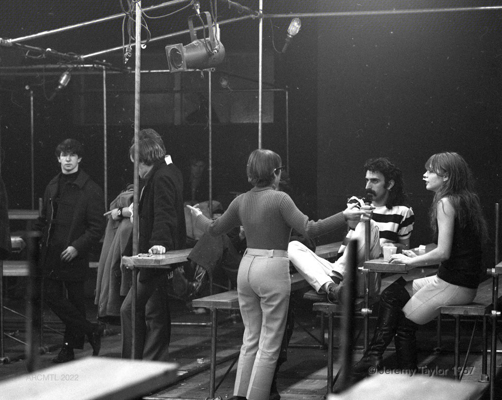 Photographie en noir et blanc de personnes debout et assises discutant dans le New Penelope. Le musicien Frank Zappa est assis sur un banc à côté d'une jeune femme assise sur le côté gauche de l'image, tout en écoutant une femme aux bras écartés qui se tient devant lui.