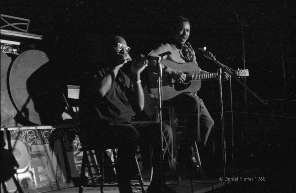 Photographie en noir et blanc des artistes Sonny Terry et Brownie McGhee assis sur des chaises au centre d'une scène, l'un jouant de la guitare et chantant, l'autre de l'harmonica, derrière des microphones. L'espace est faiblement éclairé.