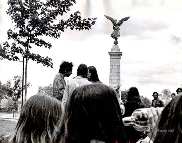 Image en noir et blanc de dix jeunes gens debout et assis dans un parc autour d'une statue au centre de l'image. La statue représente un ange ailé en pierre sur un haut et étroit piédestal en pierre.