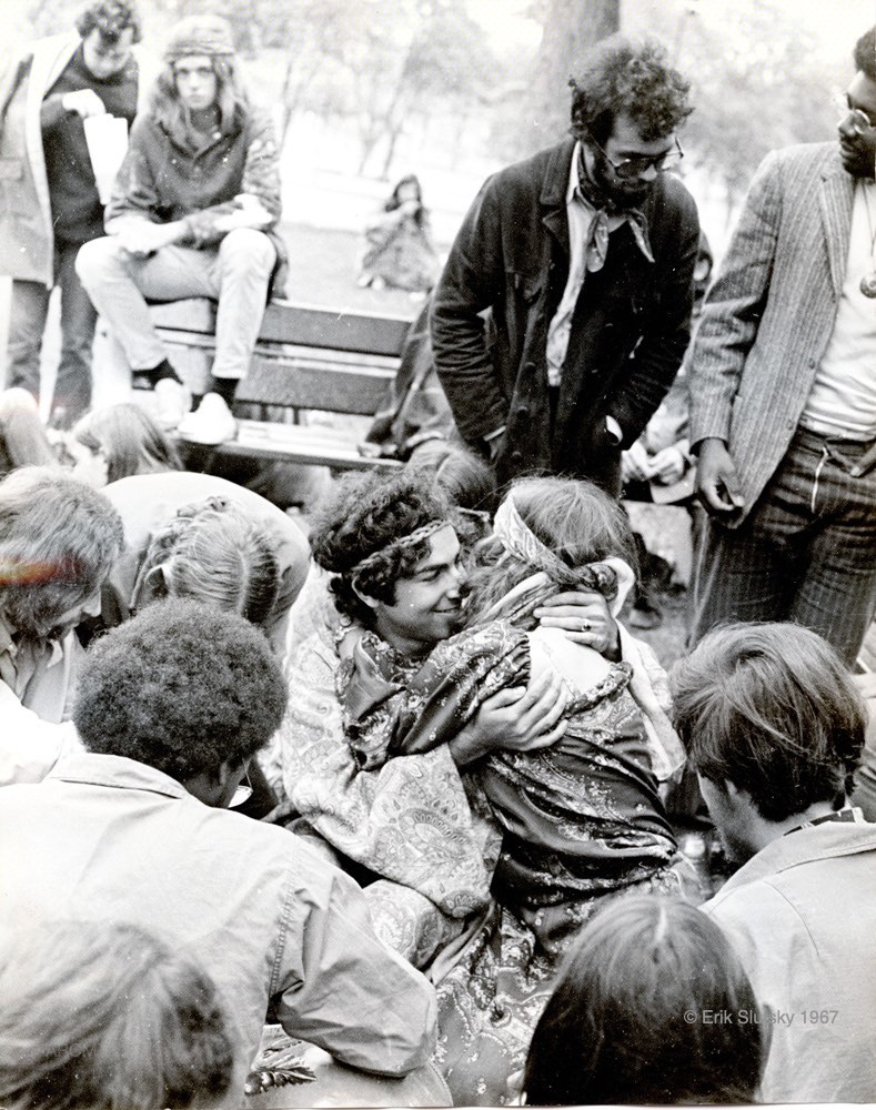 Photographie en noir et blanc de plusieurs jeunes gens assis sur des bancs dans un parc ainsi que sur le sol, avec un couple assis sur le sol s'étreignant au centre de l'image.