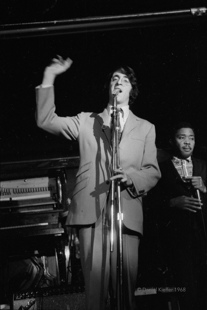 Un homme de grande taille portant des lunettes, un costume et une cravate de couleur pâle se tient devant un microphone et fait une annonce sur la scène du New Penelope, avec un musicien visible derrière lui d'un côté et un piano de l'autre