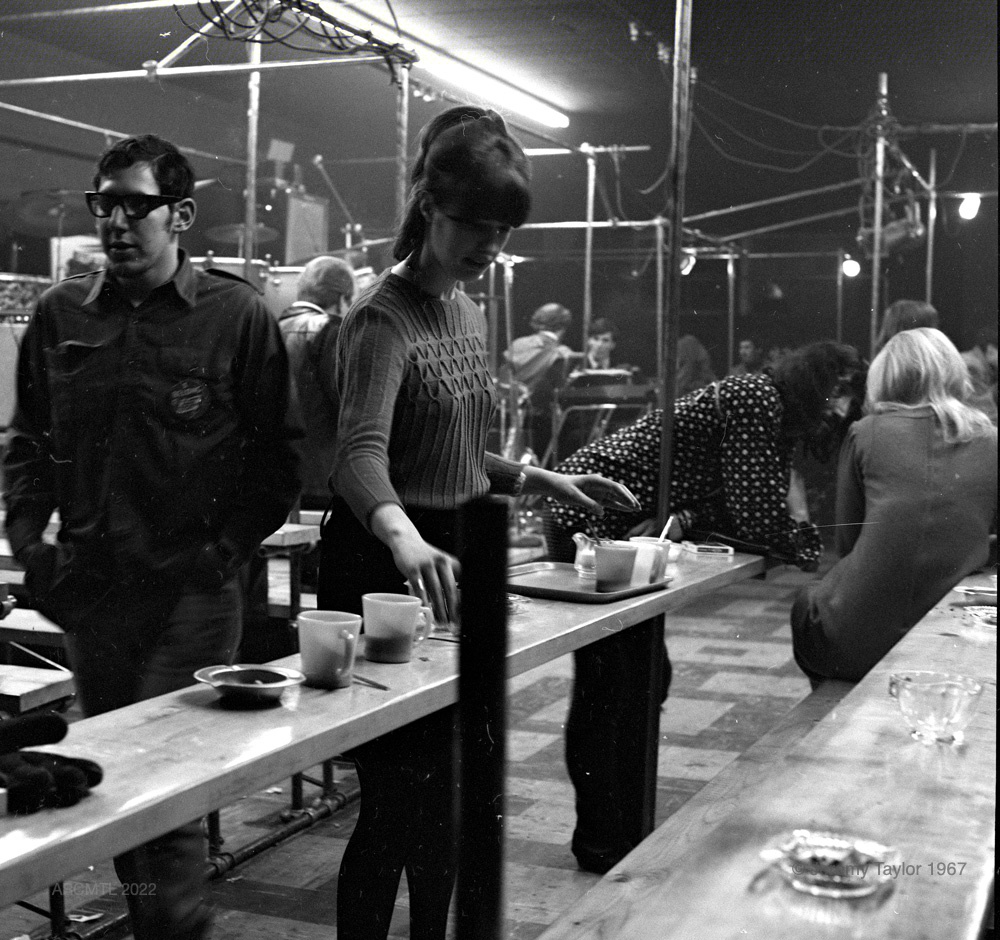 Photographie carrée en noir et blanc montrant un espace bien éclairé où plusieurs groupes de personnes se côtoient sur de longs bancs en bois. Au premier plan, une jeune femme vide un cendrier