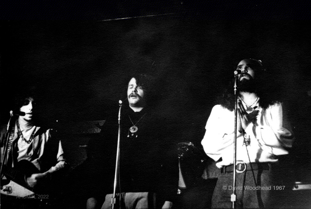 Photographie en noir et blanc représentant trois membres du groupe The Fugs chantant dans des microphones sur la scène du New Penelope, dans l'obscurité