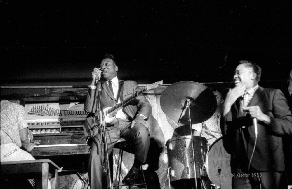 Photographie en noir et blanc représentant le chanteur de blues Muddy Waters et sa guitare, assis au centre de la scène, vêtu d'un costume et d'une cravate, avec son harmoniciste à sa gauche et son pianiste à sa droite, se produisant dans la salle de concert The New Penelope