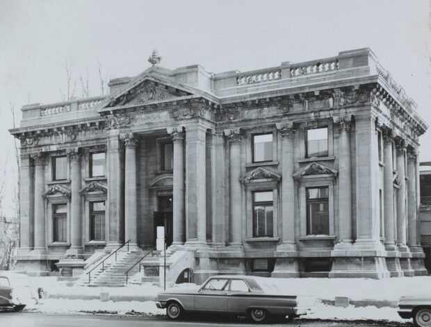 Photographie en noir et blanc d’un paysage urbain d’hiver. Le sujet de l’image au centre est une grande bâtisse d’inspirations beaux-arts faite de grandes briques de pierres grises. En avant-plan, on voit la rue et quelques voitures stationnées. 