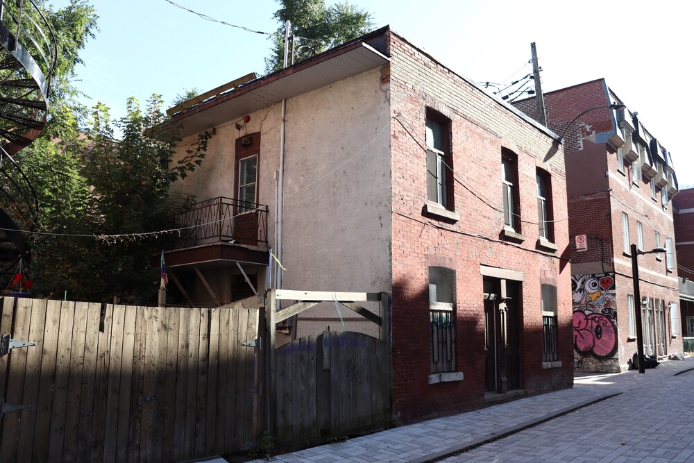 Photographie en couleur d’une ruelle sur laquelle se trouve plusieurs arrières de bâtiments. Au centre, le sujet est un petit duplex à la façade de briques orangées. Sa porte donne à même la ruelle et il est bordé d’une clôture de bois. 