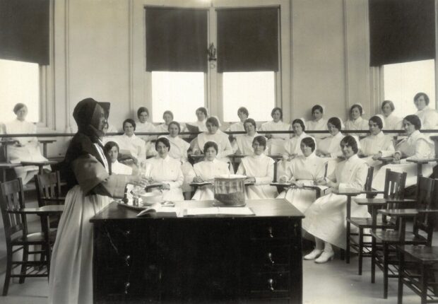 Photographie en noir et blanc d’une salle de forme ronde ou octogonale avec des fenêtres sur chaques murs et un plafond haut. La salle est organisée en estrade de trois étages. Une vingtaine d’étudiantes gardes-malades sont assises sur des chaises-bureaux. Elles portent des habits d’infirmières. En avant-plan, une femme debout en habit de religieuse semblent manipuler des récipients et verser des liquides.