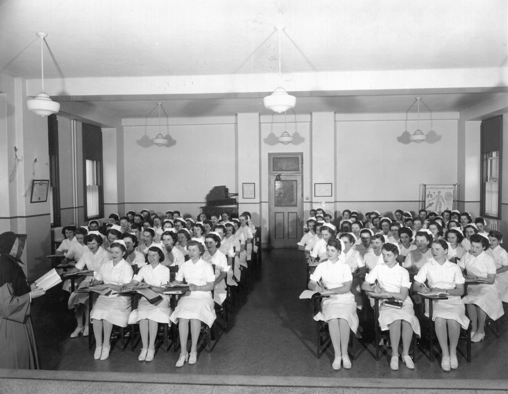 Photographie en noir et blanc d’une grande salle de classes. Des fenêtres bordent les murs des côtés, la porte est au centre du mur du fond et une affiche d'anatomie est installée au fond. En avant-plan, une femme debout en habit de religieuse tient un livre ouvert. Plus d’une soixantaine d’étudiantes en habits d’infirmières sont assises à des chaises-bureaux et prennent des notes. 