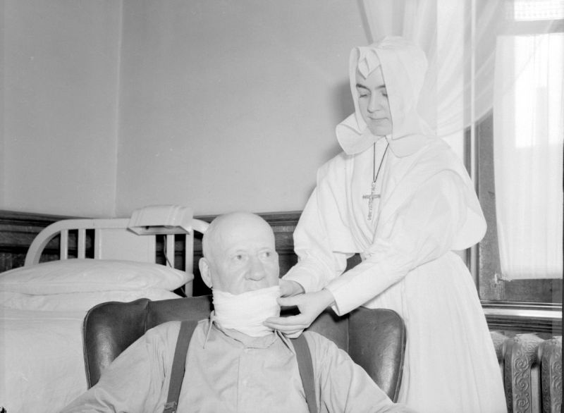 Photographie en noir et blanc d’une femme en habit de religieuse et d’un patient. Elle est debout derrière son patient qui est assis. Elle est en train de changer son pansement situé au niveau du cou et du bas du visage. 