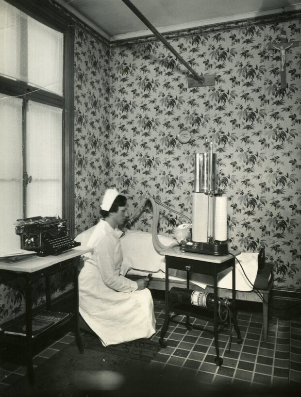 Photographie en noir et blanc d’une petite salle aux murs de papier peint fleuri. Un lit, une chaise et une petite table avec une machine à écrire meublent l’espace. Un.e patient.e impossible à identifier est couché sur le lit. Une femme en habit d’infirmière semblent manoeuvrer un tube relié a une machine vers le visage de la patient.e.