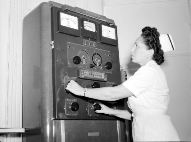 Photographie en noir et blanc d’une femme avec son habit d’infirmière. Elle est en train de manipuler des boutons rotatifs sur une grande machine rectangulaire qui affiche plusieurs cadrans.