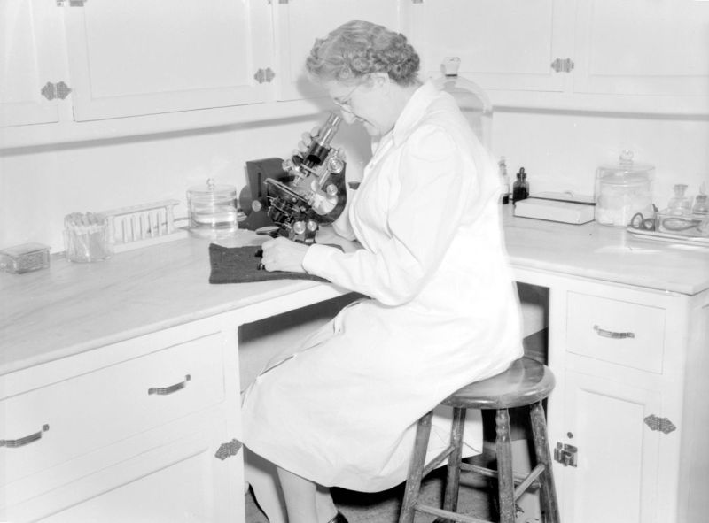 Photographie en noir et blanc prise dans une pièce qui ressemble à un laboratoire. Une femme en sarreau est assise sur un banc et regarde à travers un microscope.