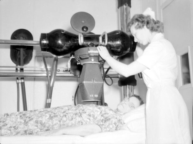  Photographie en noir et blanc représentant une petite pièce fermée peinte en blanc. Couchée sur le dos sur un lit d’hôpital, une femme reçoit un traitement d’une machine visée vers son torse. Celle-ci est manipulée par une seconde femme en habit d’infirmière. Un grand support en métal est installé dans la pièce et permet le mouvement de la machine qui ressemble à un très grand objectif de caméra.